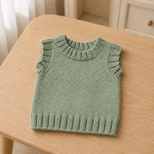 Pode incluir: Um colete de malha sem mangas na cor verde sálvia, com gola redonda canelada, cavas caneladas com um detalhe subtil de folhos e uma bainha larga canelada. A peça é confecionada com um padrão clássico de ponto jersey em todo o corpo.