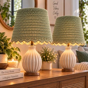 May include: Two table lamps featuring white ceramic bases with vertical ribbed textures and round wooden stands. Each lamp is topped with a sage green crocheted lampshade that has a scalloped edge, emitting a warm glow from within the shade.