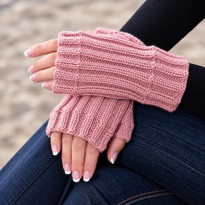 May include: A pair of dusty rose fingerless gloves featuring a vertical ribbed knit pattern. The gloves cover the hand from the knuckles to the wrist, showcasing a textured, knitted construction with a consistent color throughout the fabric.