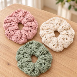 May include: Three circular hair scrunchies made of thick, textured yarn. One scrunchie is dusty pink, one is cream, and one is sage green. Each item features a gathered, crocheted design with a central opening for securing hair.