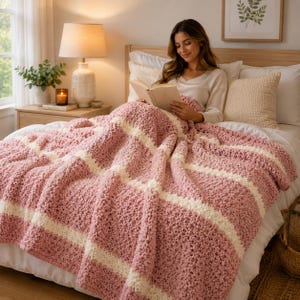 May include: A chunky knit throw blanket featuring a repeating pattern of wide dusty rose pink stripes and narrower cream white stripes. The blanket has a soft, textured, looped yarn construction and is draped over a bed with white bedding.