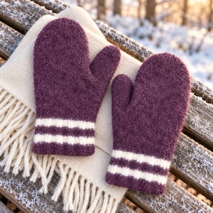 May include: A pair of dark purple wool mittens featuring two horizontal white stripes near the wrist, resting on a folded cream-coloured knit scarf with fringe detailing at the bottom edge, showcasing a textured and thick material construction.