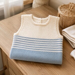 Pode incluir: Um colete de malha sem mangas com uma secção superior em cor creme e uma secção inferior com riscas horizontais alternadas em creme e azul claro. A peça tem um decote redondo com um fecho de botão único no centro da gola.