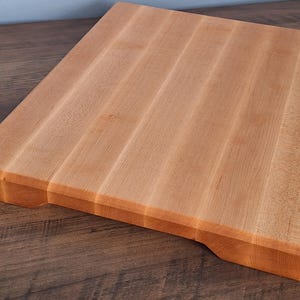 May include: A rectangular wooden chopping board made of light-coloured natural wood with a smooth, flat surface and visible grain patterns. The board features thick edges and small, carved indentations on the underside for easy lifting and handling.