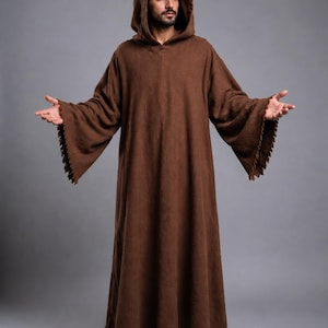 May include: A floor-length brown hooded robe featuring long, wide sleeves with a fringed edge. The garment is made of a textured fabric and includes a hood that rests over the shoulders. The hem of the robe also features a matching fringed detail.
