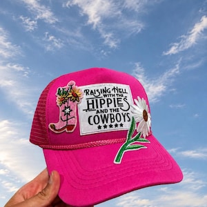 Może przedstawiać: Jaskraworóżowa czapka typu trucker z białym prostokątnym naszywką z czarnym napisem "RAISING HELL WITH THE HIPPIES AND THE COWBOYS" oraz pięcioma gwiazdkami. Czapka jest ozdobiona wyhaftowanym różowym kowbojskim butem i białym kwiatem stokrotki.