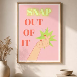 以下が含まれることがあります： ライトピンクの背景にライムグリーンの「SNAP」、コーラルレッドの「OUT OF IT」という文字が描かれた額入りアートプリント。指を鳴らすジェスチャーをする手のイラストが、ライムグリーンのスターバーストグラフィックの横に配置されています。