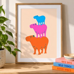 May include: A framed art print featuring a stack of three capybaras in bright colours against a light peach background. The top capybara is blue, the middle is pink, and the bottom is orange. The print is displayed in a light wood frame.