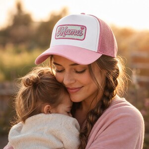 May include: A trucker hat featuring a white front panel, a pink brim, and pink mesh back panels. The front displays a rectangular patch with a glittery pink border and the word Mama in a cursive script font, accompanied by a small heart icon.