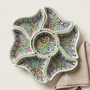 Peut inclure: Un ensemble de service en céramique composé d'un bol circulaire central entouré de six coupelles en forme de pétale. Chaque pièce présente un motif géométrique peint à la main dans des tons vifs de vert, rouge, bleu et orange sur fond blanc.