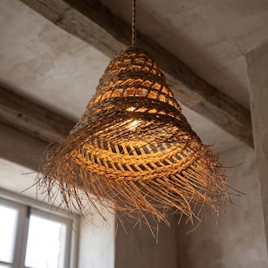 May include: A bell-shaped pendant light fixture crafted from woven natural fibers in a light tan color. The structure features a lattice pattern at the top that transitions into a frayed, textured fringe at the bottom edge, emitting a warm glow.