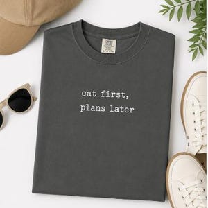 Peut inclure: Un t-shirt à col rond gris anthracite avec le texte blanc "cat first, plans later" imprimé sur la poitrine. Le t-shirt présente une étiquette de marque visible à l'intérieur du col et est accompagné d'une casquette beige, de lunettes et de baskets blanches.