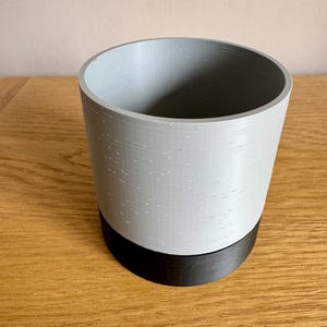 May include: A cylindrical planter featuring a two-tone design with a light grey upper section and a solid black base. The exterior surface displays a subtle horizontal texture characteristic of 3D printed construction, forming a simple open vessel.