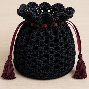 May include: A black crochet drawstring pouch featuring an openwork lattice pattern, a scalloped top edge, and a deep burgundy ribbon closure threaded through the upper section, finished with two matching burgundy tassels at the ends.