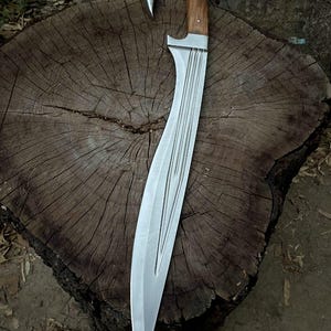 May include: A curved steel blade with a sharp point and a series of parallel grooves running along the center. The handle is crafted from polished brown wood with a distinct hook-shaped pommel and two small metal pins securing the grip.