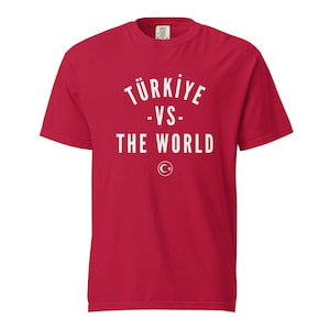 Puede incluir: Una camiseta roja de manga corta con texto blanco que dice TÜRKIYE -VS- THE WORLD en un diseño apilado. Debajo del texto hay un pequeño emblema circular blanco que contiene una luna creciente y una estrella de cinco puntas en el pecho.