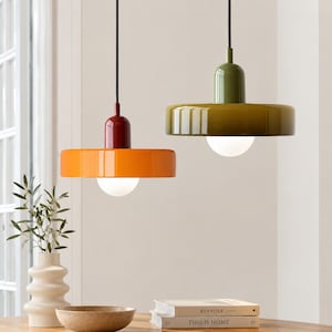 Op de afbeelding: Twee hanglampen hangen aan zwarte snoeren. De ene lamp heeft een glanzende oranje kap met een donkerrode dop, terwijl de andere een glanzende olijfgroene kap heeft. Beide lampen bevatten een ronde witte lamp onder de platte kappen.