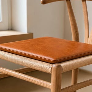 Puede incluir: Un cojín de asiento de cuero marrón claro colocado sobre una silla de madera con asiento de cuerda trenzada. El cojín es rectangular con esquinas redondeadas y una superficie lisa, apoyado directamente sobre la base trenzada de la silla.