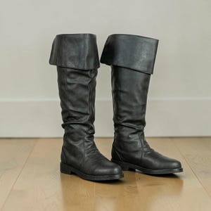 May include: A pair of black knee-high boots featuring a wide, folded cuff at the top. The boots have a smooth, matte finish, a rounded toe, and a low, flat heel. The shaft of the boots shows subtle vertical stitching and a slightly slouchy texture.