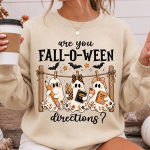 Op de afbeelding: Een beige sweater met ronde hals en de zwarte tekst "are you FALL-O-WEEN directions?" met een afbeelding van drie geesten aan een waslijn. De geesten houden een potlood, een boek met "LESSONS" en een notitieboek met "TEACHER NOTES" vast.