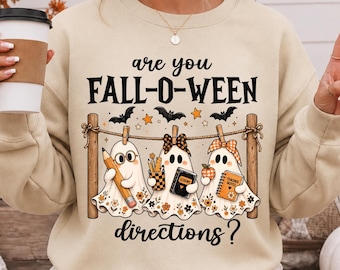 May include: A beige crewneck sweatshirt featuring the black text "are you FALL-O-WEEN directions?" and a graphic of three ghosts hanging from a clothesline. The ghosts are holding a pencil, a book labelled "LESSONS", and a notebook labelled "TEACHER NOTES".