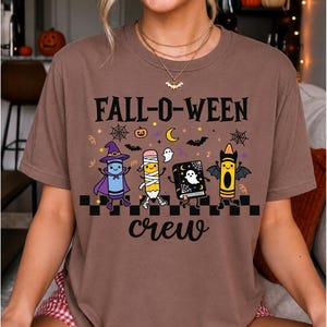 Op de afbeelding: Een bruin t-shirt met korte mouwen en de tekst FALL-O-WEEN crew in het zwart. De afbeelding toont een paars kleurpotlood verkleed als heks, een potlood als mummie, een zwart boek met een geest en een oranje kleurpotlood met vleermuisvleugels.
