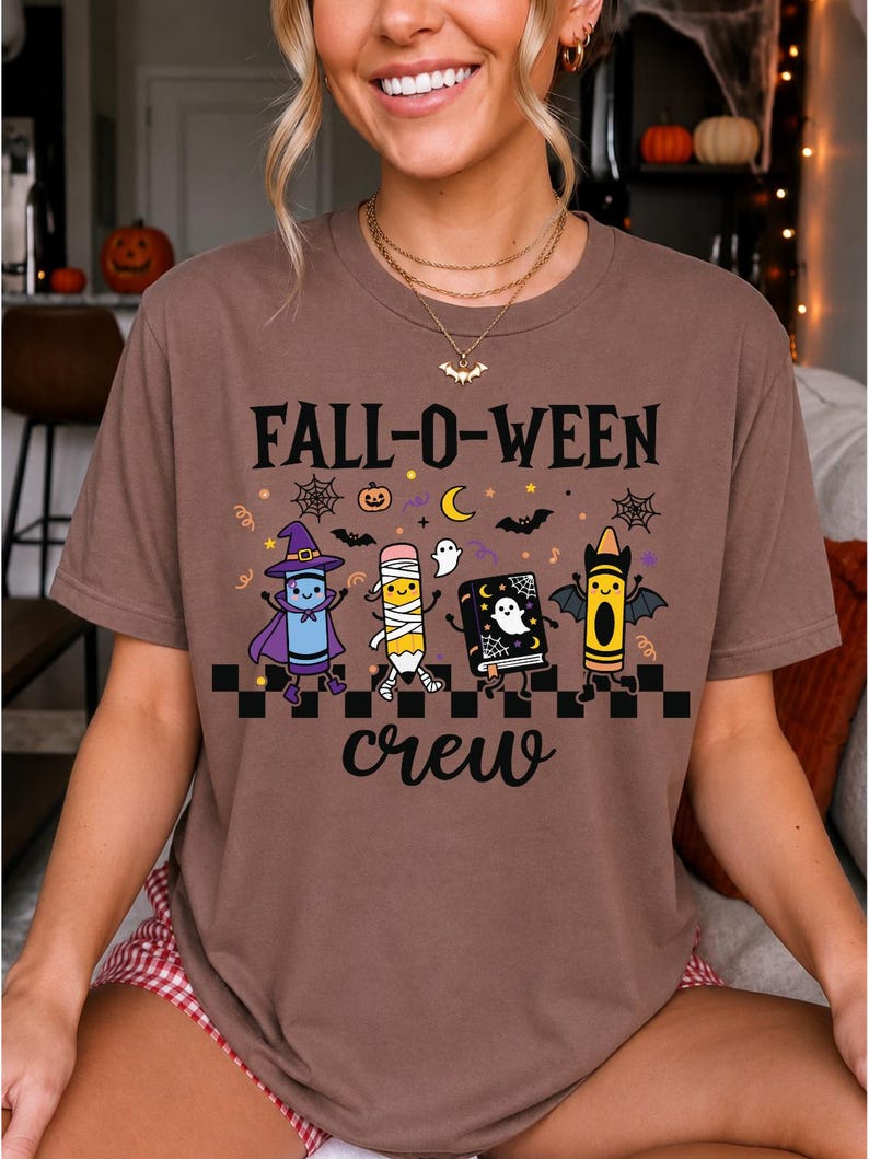Op de afbeelding: Een bruin t-shirt met korte mouwen en de tekst FALL-O-WEEN crew in het zwart. De afbeelding toont een paars kleurpotlood verkleed als heks, een potlood als mummie, een zwart boek met een geest en een oranje kleurpotlood met vleermuisvleugels.
