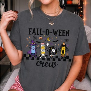 Op de afbeelding: Een antracietgrijs t-shirt met korte mouwen en de tekst FALL-O-WEEN crew in het zwart. De afbeelding toont een paars krijtje als heks, een potlood als mummie, een zwart spreukenboek met een geest en een geel krijtje als vleermuis.