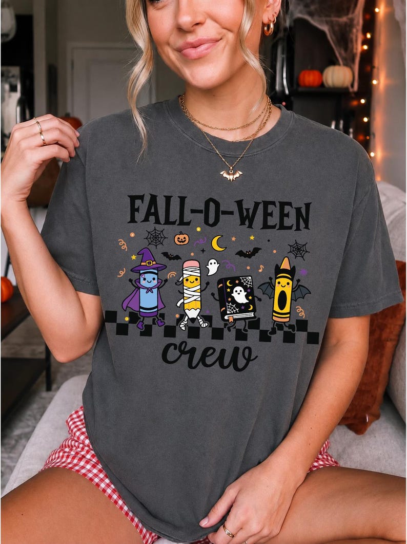 Op de afbeelding: Een antracietgrijs t-shirt met korte mouwen en de tekst FALL-O-WEEN crew in het zwart. De afbeelding toont een paars krijtje als heks, een potlood als mummie, een zwart spreukenboek met een geest en een geel krijtje als vleermuis.