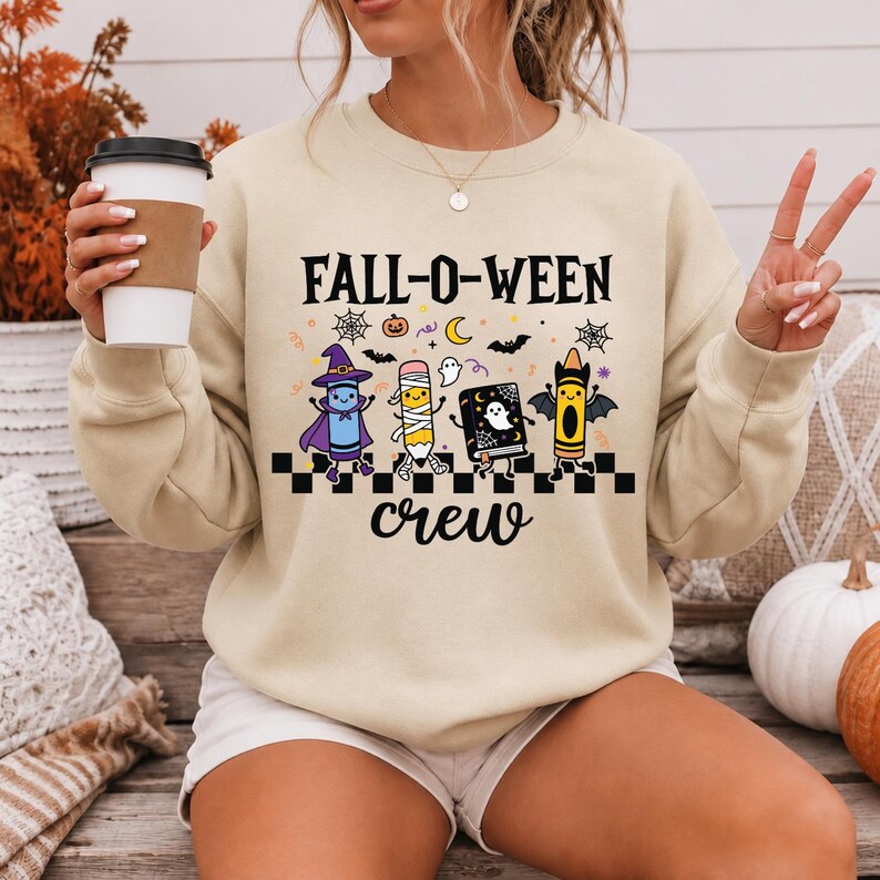 Op de afbeelding: Een beige sweater met ronde hals en de zwarte tekst FALL-O-WEEN crew. De afbeelding toont een paars kleurpotlood verkleed als heks, een potlood als mummie, een zwart boek met een geest en een geel kleurpotlood met vleermuisvleugels boven een dambord.