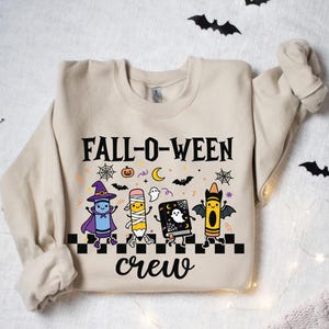 Op de afbeelding: Een beige sweater met ronde hals en een grafische print met de tekst FALL-O-WEEN crew. Het ontwerp bevat een blauw kleurpotlood verkleed als heks, een potlood verkleed als mummie, een boek met een geest en een geel kleurpotlood met vleermuisvleugels.