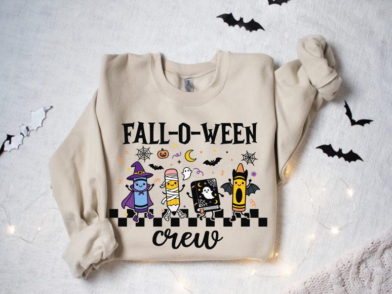 Op de afbeelding: Een beige sweater met ronde hals en een grafische print met de tekst FALL-O-WEEN crew. Het ontwerp bevat een blauw kleurpotlood verkleed als heks, een potlood verkleed als mummie, een boek met een geest en een geel kleurpotlood met vleermuisvleugels.