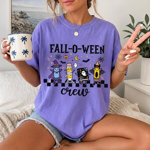 Op de afbeelding: Een paars t-shirt met de tekst FALL-O-WEEN crew en een afbeelding van vier stripfiguren, waaronder een kleurpotlood verkleed als heks, een potloodmummie, een boek met een geest en een kleurpotlood-vleermuis, boven een zwart-wit geblokte rand.