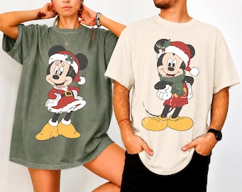 Op de afbeelding: Een donker olijfgroen t-shirt met een afbeelding van Minnie Mouse in een kerstmuts en jurk, en een crèmekleurig t-shirt met een afbeelding van Mickey Mouse in een kerstmuts en sjaal. Beide shirts hebben een verweerde vintage-stijl print.