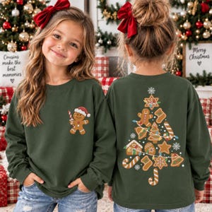 May include: A dark green long-sleeve sweatshirt featuring a small gingerbread man graphic on the front left chest and a large graphic on the back depicting a Christmas tree composed of various gingerbread cookies, candy canes, and snowflakes.
