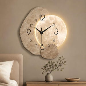 Può includere: Un orologio da parete con un quadrante circolare illuminato simile alla luna, con numeri e lancette neri, su uno sfondo irregolare color beige con texture in pietra. La luce emette un bagliore soffuso da dietro il quadrante circolare.