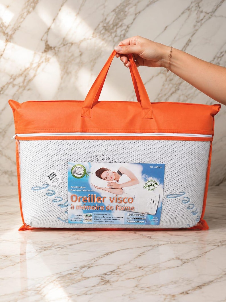 May include: A rectangular memory foam pillow packaged in a clear plastic bag with orange fabric trim and handles. The label features the text Oreiller visco &agrave; m&eacute;moire de forme, 40 x 60 cm, and an illustration of a person sleeping on a pillow.