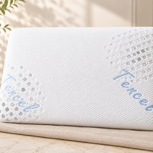 May include: A rectangular white pillow with a textured knit surface featuring two circular patterns of grey dots. The word Tencel is embroidered in light blue script on the left and right sides of the pillow, positioned over the grey dot patterns.