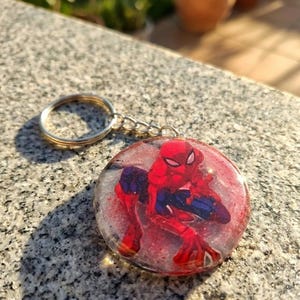 Può includere: Un portachiavi rotondo con un design di Spider-Man. L'immagine di Spider-Man è in rosso e blu, su uno sfondo rosso e glitterato. Il portachiavi ha un anello e una catena color argento.