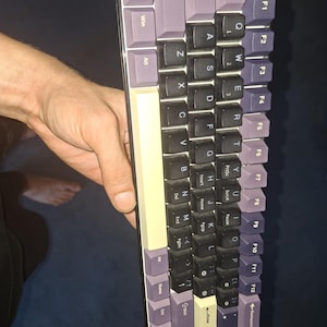 Könnte beinhalten: Eine kompakte mechanische Tastatur mit einer Farbgestaltung aus schwarzen, hellvioletten und cremefarbenen Tastenkappen. Das Layout umfasst standardmäßige alphanumerische Tasten in Schwarz, Modifikatortasten in Hellviolett und eine cremefarbene Leertaste.