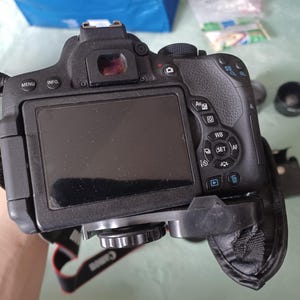 May include: A black digital SLR camera featuring a large rear LCD screen, a viewfinder, and various control buttons including Menu, Info, Av, WB, and Set. The camera body has a textured grip and is attached to a black fabric neck strap with Canon branding.