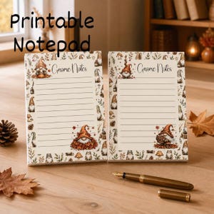 May include: Two rectangular notepads with cream-colored paper featuring horizontal lines and the text Gnome Notes. The borders are decorated with illustrations of gnomes, autumn leaves, mushrooms, and pinecones. A gold fountain pen lies nearby.