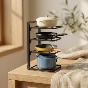 May include: A black metal four-tier storage rack holding a cream-colored lidded pot, a black frying pan, a yellow frying pan with a wooden handle, and a blue pot with two side handles, arranged vertically on a wooden surface.