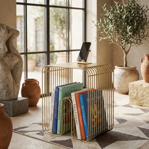 May include: A gold-toned metal side table with a wire frame design, featuring a flat top surface holding a black smartphone stand. The base of the table serves as a storage rack for several books with spines labeled Art de Vivre and Déco Française.