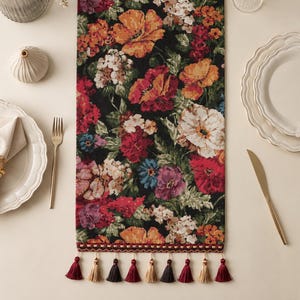 May include: A rectangular table runner featuring a dense, colorful floral pattern on a black background. The design includes red, orange, white, and blue flowers with green foliage. The bottom edge is finished with a decorative trim and tassels.