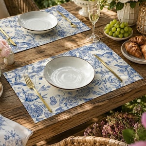 May include: Two rectangular placemats featuring a blue and white floral toile pattern. Each placemat is set with a white ceramic bowl, a gold-toned fork, and a gold-toned knife. The placemats are arranged on a rustic wooden table with place settings.