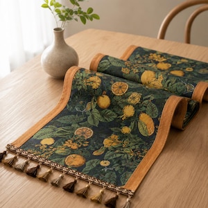 Puede incluir: Un camino de mesa rectangular con fondo verde oscuro y un patrón repetitivo de frutas cítricas amarillas y hojas verdes. El camino tiene un borde naranja sólido y está rematado con una fila de pequeñas borlas marrones en el borde.