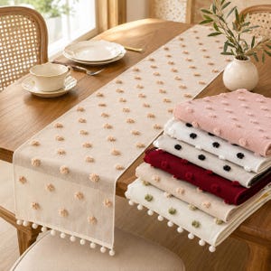 May include: A beige table runner with a repeating pattern of small tufted pom-poms and a pom-pom fringe edge. A stack of folded table runners in various colors including pink, white, black, red, and beige sits on the wooden table surface.