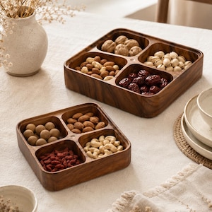 May include: Two square wooden serving trays with four divided compartments each, filled with assorted nuts and dried fruits. The trays feature a dark brown wood grain finish and are arranged on a white textured surface alongside a ceramic vase.