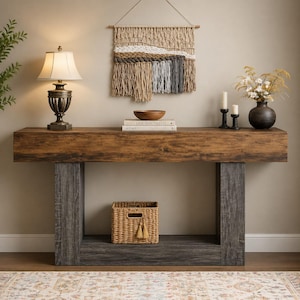 May include: A rectangular console table with a dark wood grain top and charcoal gray legs. The table features a lower shelf holding a woven basket. On top sits a lamp, a wooden bowl, two books titled Live Beautifully and Home, and a vase with flowers.